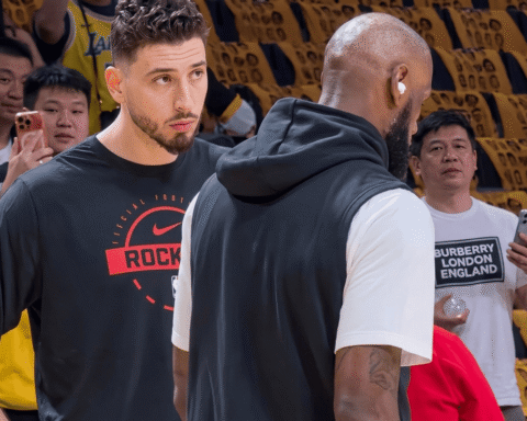 Two basketball players converse on a court; one faces the camera in a black shirt with a red logo, the other has his back turned in a white T-shirt with a vest.