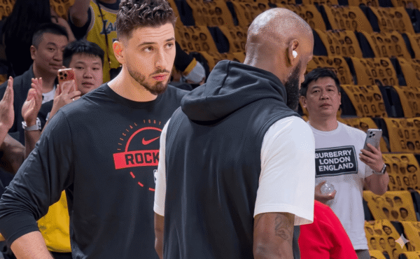 Two basketball players converse on a court; one faces the camera in a black shirt with a red logo, the other has his back turned in a white T-shirt with a vest.