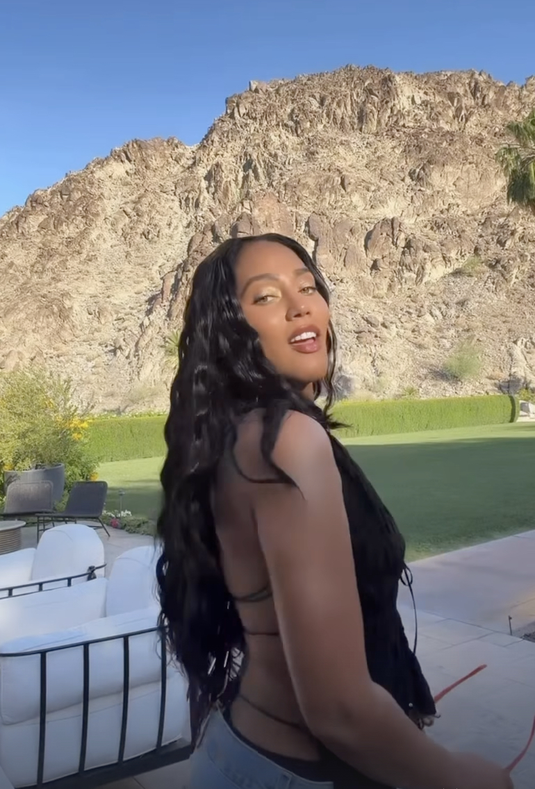 Woman with long dark hair posing outdoors, rocky hillside and blue sky behind her, wearing a black backless top and jeans.