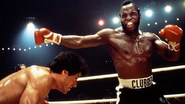 Boxer in white-trimmed shorts celebrates victory with arms raised as opponent crouches in the ring.