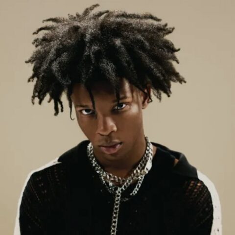 Portrait of a young man with a large afro, wearing a black knit shirt and chunky chains, gazing intently at the camera.