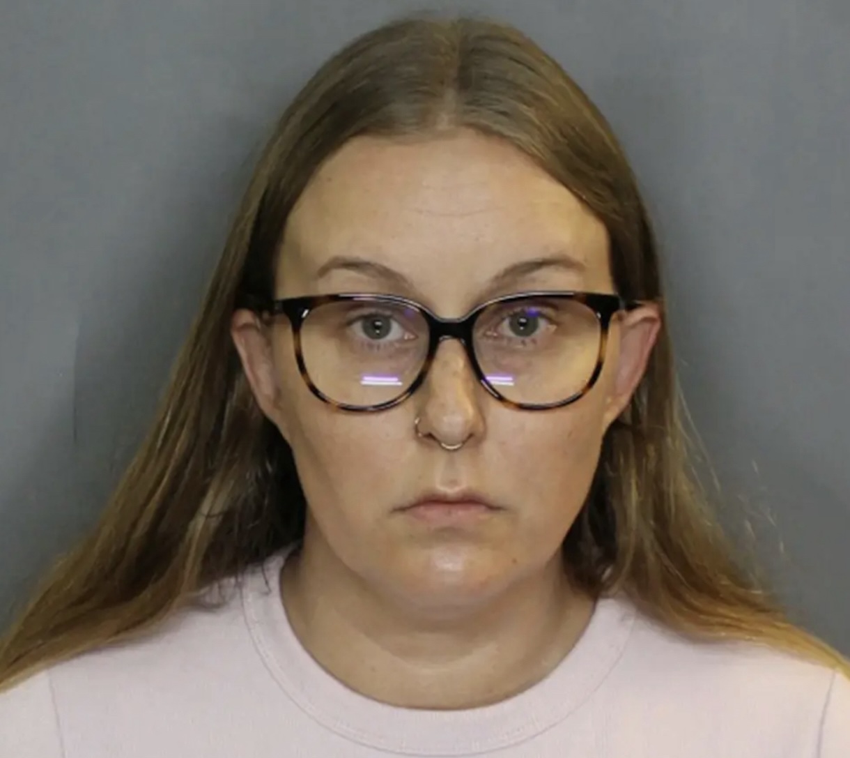 Front-facing mugshot-style portrait of a young woman with long light brown hair, wearing large tortoiseshell glasses and a nose ring, in a pale pink shirt against a gray backdrop.