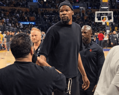 Basketball player in a black shirt standing on a court, surrounded by teammates and staff with a crowded arena in the background.