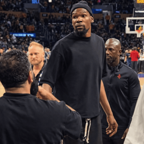 Basketball player in a black shirt standing on a court, surrounded by teammates and staff with a crowded arena in the background.