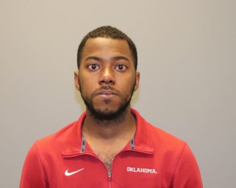 Mugshot-style photo of a man in a red Oklahoma jacket against a plain gray background.