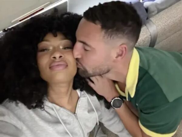 Couple lying close together; man kisses the woman's cheek as she smiles, both in casual clothing on a beige couch/bed.