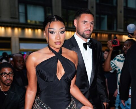 Glamorous couple in formal wear walking a red carpet; woman in a black halter gown with a front cutout and jeweled belt, man in a black tuxedo behind her.