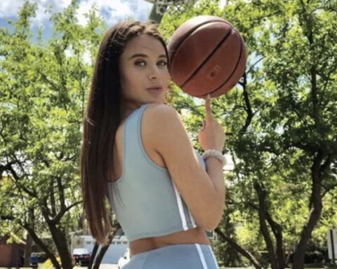 Woman in a light blue athletic outfit outdoors, spinning a basketball on her finger with trees in the background.