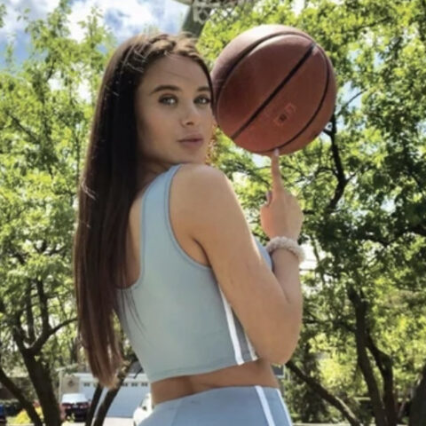 Woman in a light blue athletic outfit outdoors, spinning a basketball on her finger with trees in the background.