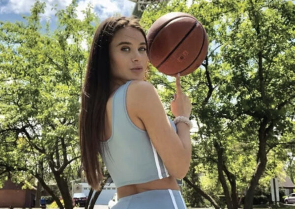 Woman in a light blue athletic outfit outdoors, spinning a basketball on her finger with trees in the background.