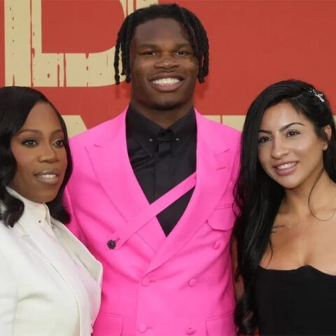 Three people posing on a red-carpet backdrop, smiling at the camera: woman in white blazer, man in a bright pink suit, woman in a black strapless dress with a colorful sleeve tattoo visible on her arm.