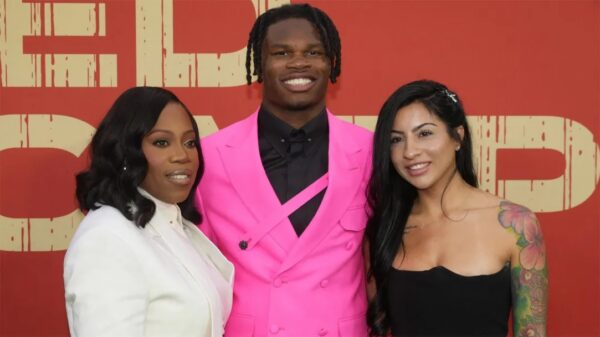 Three people posing on a red-carpet backdrop, smiling at the camera: woman in white blazer, man in a bright pink suit, woman in a black strapless dress with a colorful sleeve tattoo visible on her arm.