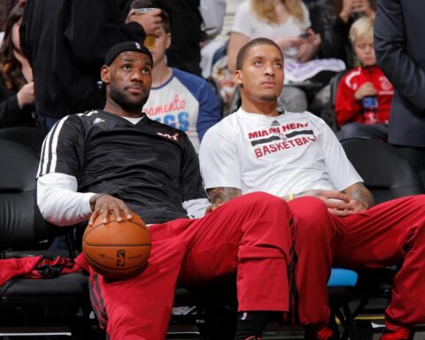 Two Miami Heat players sit courtside in team warmups; one holds a basketball and looks upward while the other gazes ahead, with fans in the background.