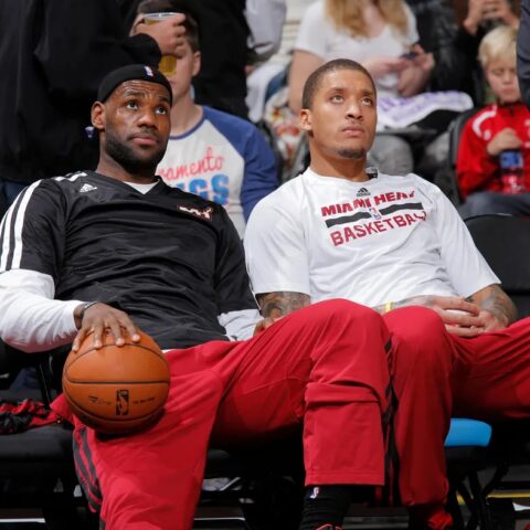 Two Miami Heat players sit courtside in team warmups; one holds a basketball and looks upward while the other gazes ahead, with fans in the background.