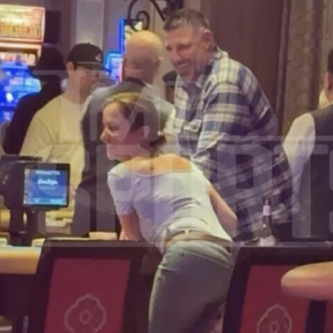 Woman leaning on a casino bar, smiling, with a tall man in a plaid shirt beside her and others in the background.