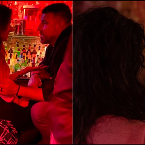 Two people sit at a red-lit bar, facing each other and holding hands in a close, intimate conversation at nightlife venue.