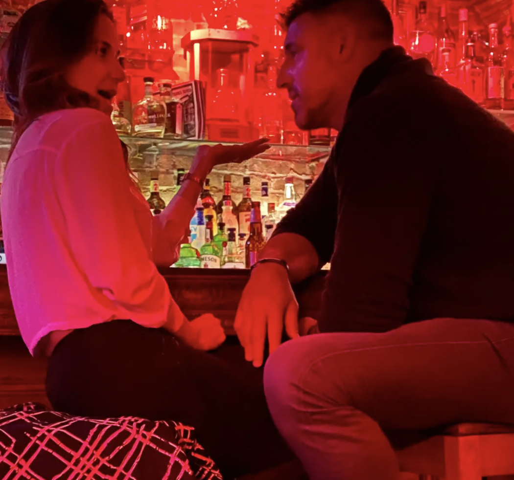 Two people chatting at a dimly lit bar, facing each other and resting elbows on the counter under red lighting.