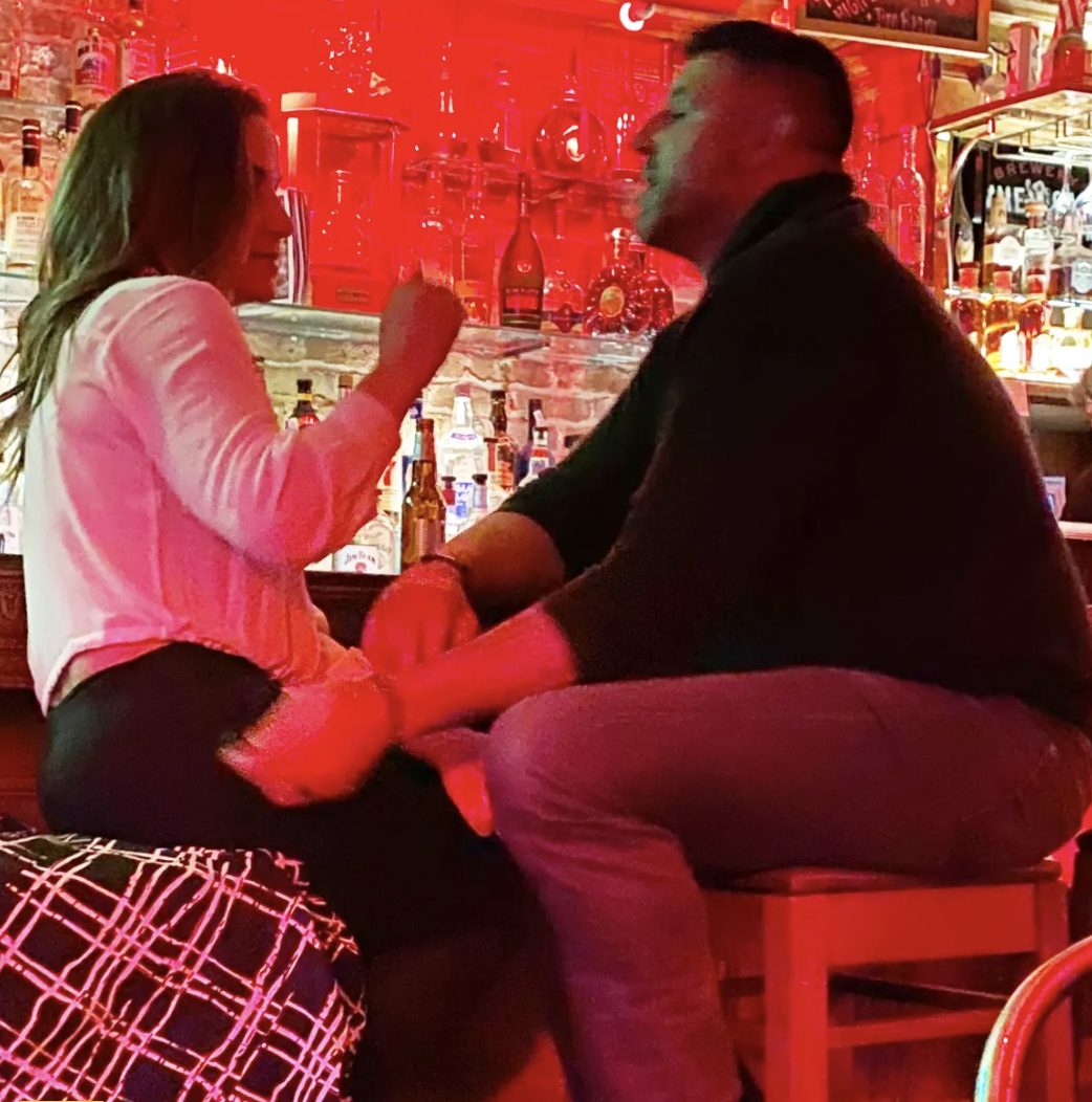 Two people sit on bar stools at a dimly lit bar, facing each other and talking with red lighting behind them.
