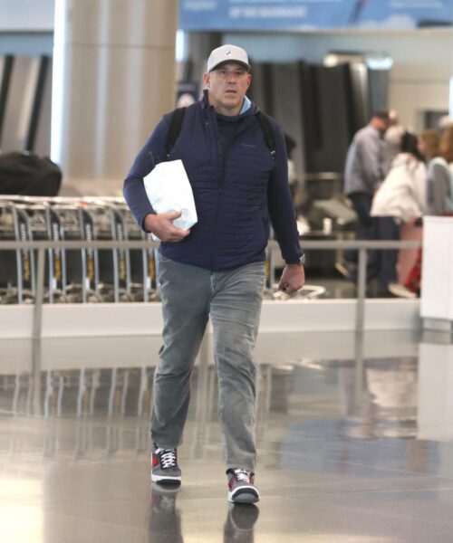 Man in a dark blue quilted vest and light gray cap walking through a busy airport, carrying a white bag and wearing a backpack.