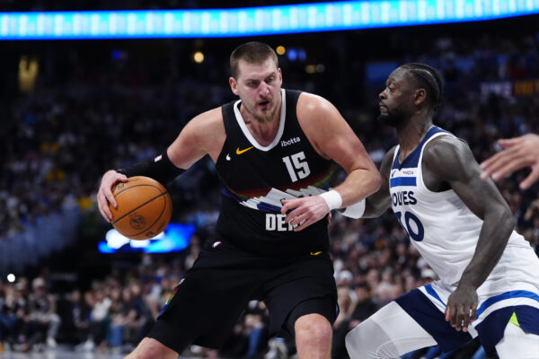 Two basketball players sprint on the court; black jersey #15 drives with the ball while a defender in a white Wolves jersey pursues him.