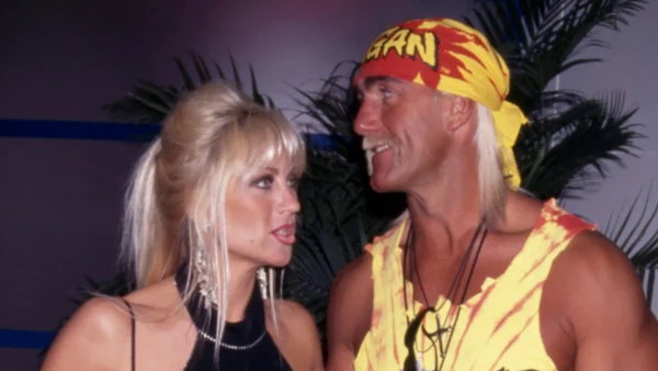 Two adults facing each other and smiling at a tropical-themed party, woman in black dress and earrings, man in yellow-red bandana and tie-dye outfit.