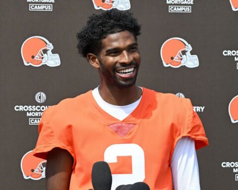 Smiling African American football player in an orange jersey (#3) at a press event with Browns logos in the background, microphones in front of him.