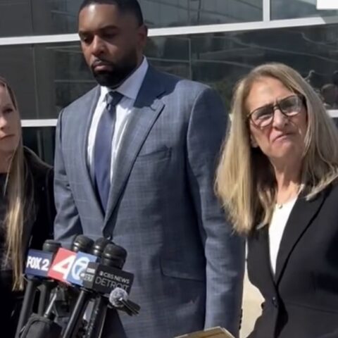 Three adults stand behind a cluster of microphones during a press conference outside a glass-front building; man in a blue suit center, two women flanking him.