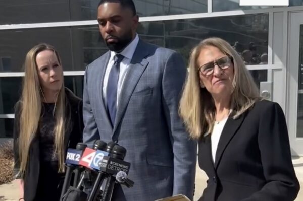 Three adults stand behind a cluster of microphones during a press conference outside a glass-front building; man in a blue suit center, two women flanking him.