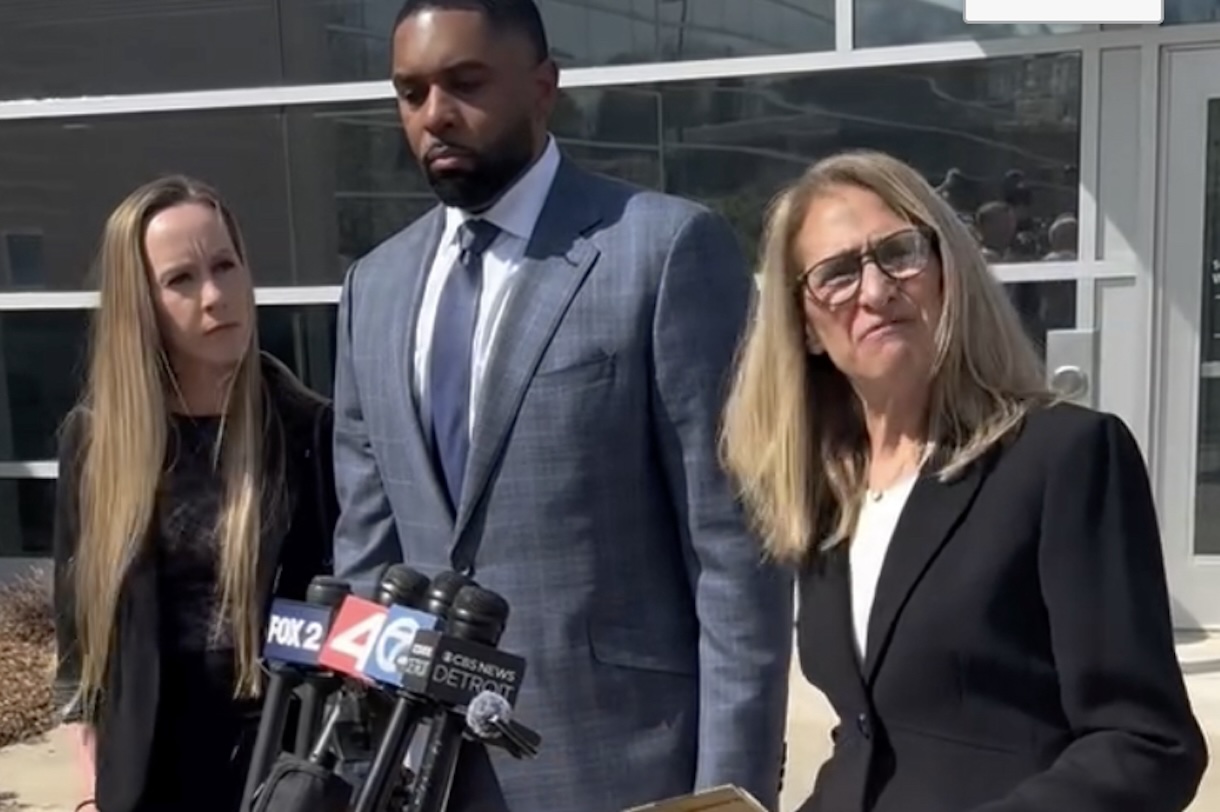 Three adults stand behind a cluster of microphones during a press conference outside a glass-front building; man in a blue suit center, two women flanking him.