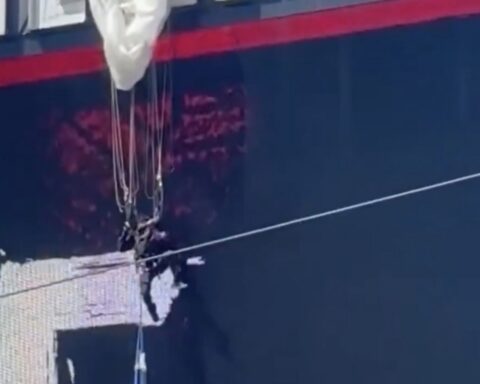 Side of a dark blue ship hull with a red stripe near the top and tangled wires hanging from a damaged area with white patches.