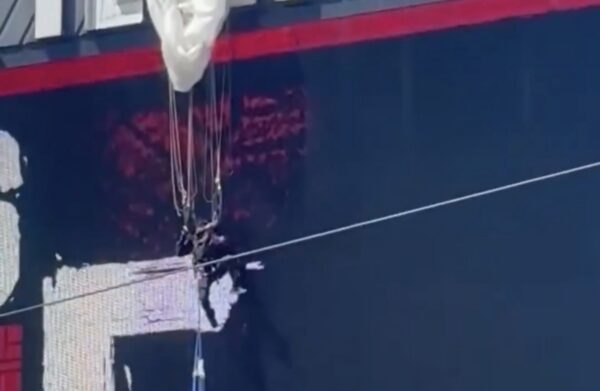 Side of a dark blue ship hull with a red stripe near the top and tangled wires hanging from a damaged area with white patches.