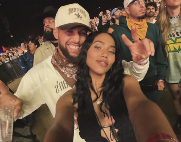 Smiling couple at a nighttime outdoor concert taking a selfie; man wears a beige cap and bandana, woman in a black lace-up top, posing for the camera with a crowd behind them.
