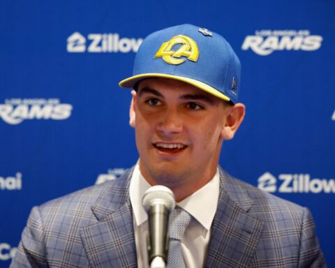 Man in a blue plaid suit speaking at a press event, wearing a blue cap with a yellow LA logo in front of a blue backdrop with sponsor logos