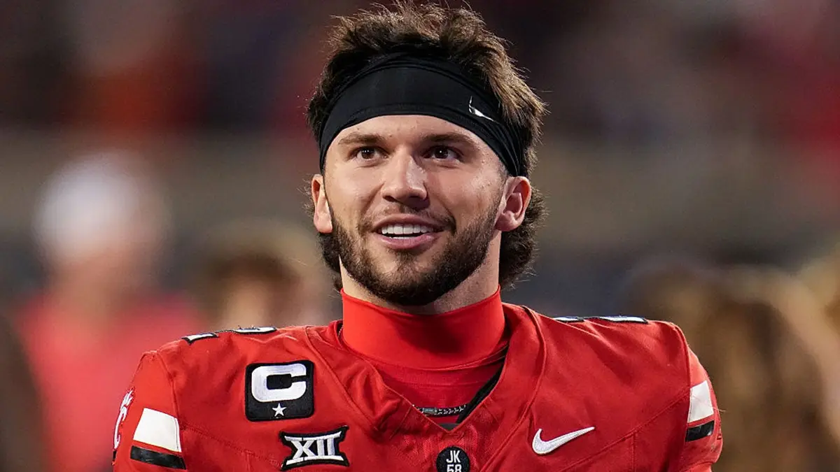 Male football player in a red jersey with captain’s patch, smiling, wearing a black headband on a blurred stadium background, likely a team captain.