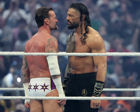 Two muscular wrestlers face off in a ring, locking eyes as a blurred crowd watches in the background.