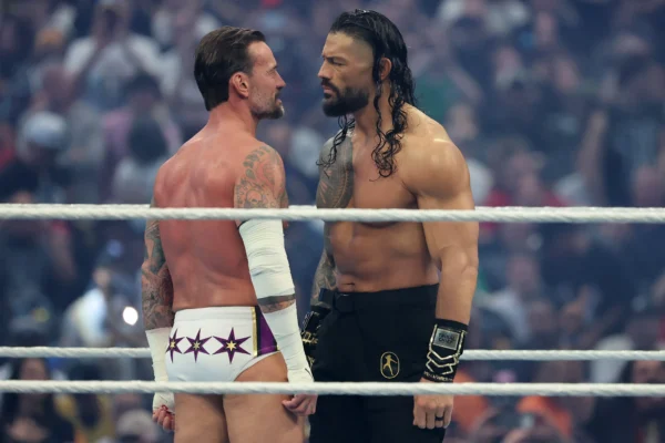 Two muscular wrestlers face off in a ring, locking eyes as a blurred crowd watches in the background.