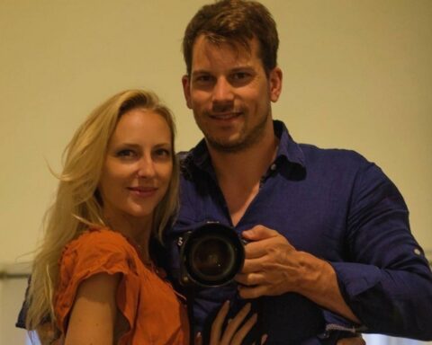 Smiling couple posing for a mirror selfie; the man holds a camera between them on a beige wall. (informative)