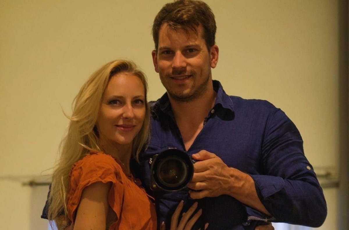 Smiling couple posing for a mirror selfie; the man holds a camera between them on a beige wall. (informative)
