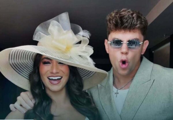 Woman wearing a large striped sun hat with a flower, smiling beside a man in a light blazer and sunglasses making a surprised expression.