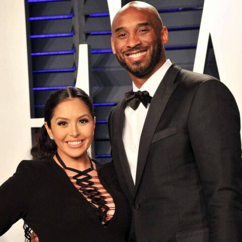 Smiling couple in formal wear posing on a red carpet; woman in a black lace-up dress and man in a black tuxedo.