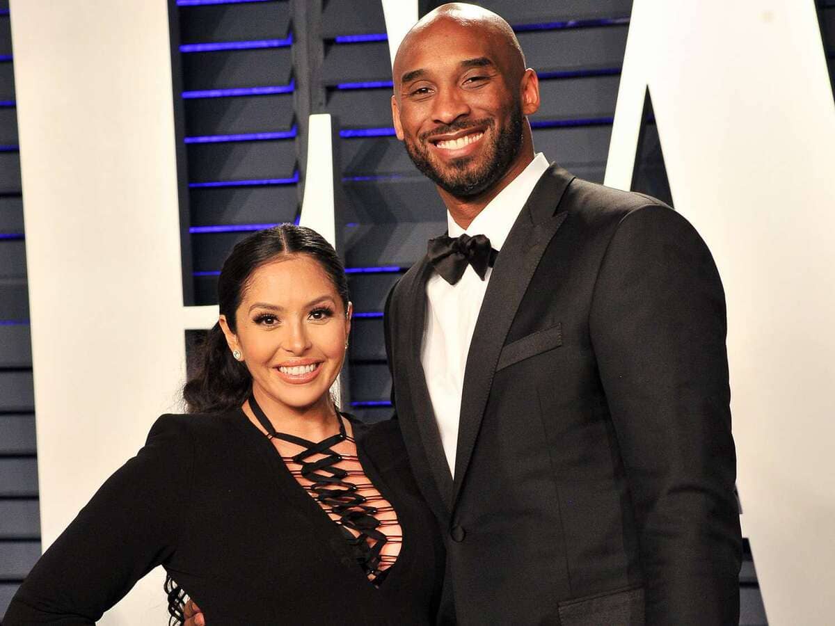 Smiling couple in formal wear posing on a red carpet; woman in a black lace-up dress and man in a black tuxedo.