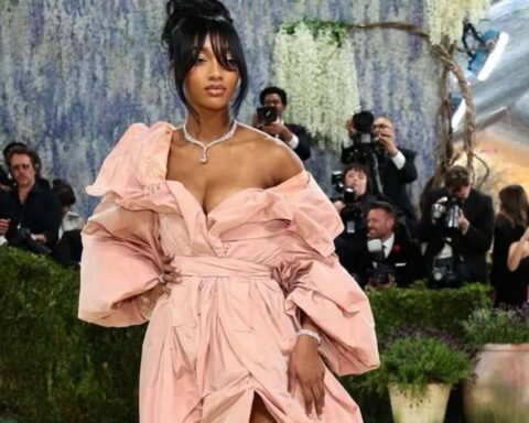 Rihanna wearing a pale pink off-the-shoulder gown with voluminous sleeves on a red carpet, posing with one hand on her hip as photographers look on.