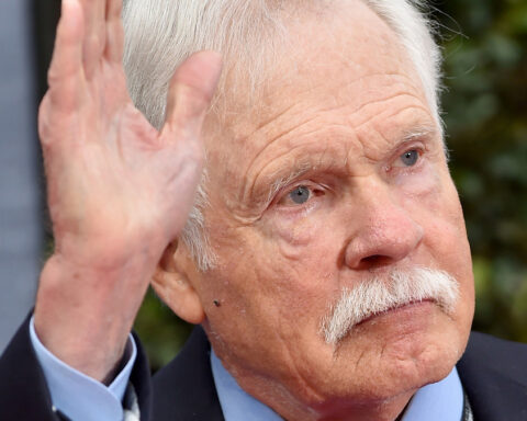 Close-up of an elderly man with a white mustache raising his right hand in a wave outdoors.
