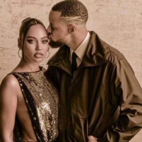 Couple posing closely; man kisses woman's temple while she looks at the camera, wearing a gold sequin dress and large hoops against a beige textured wall