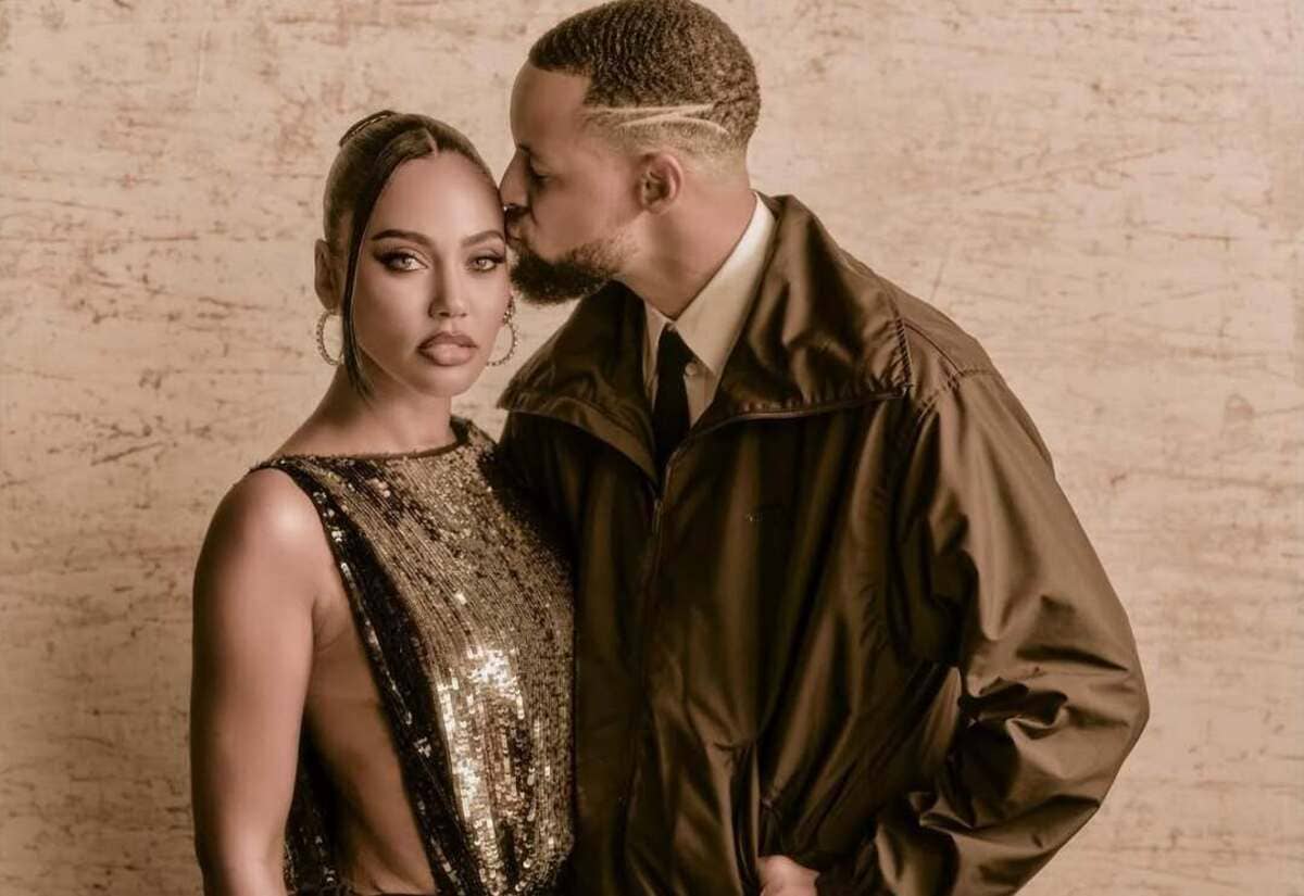 Couple posing closely; man kisses woman's temple while she looks at the camera, wearing a gold sequin dress and large hoops against a beige textured wall