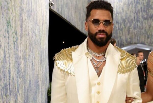 Man wearing a cream suit with gold epaulettes, multiple necklaces, and sunglasses at a formal event