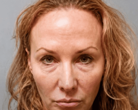 Mugshot-style portrait of a woman with reddish hair and a neutral expression against a gray background.