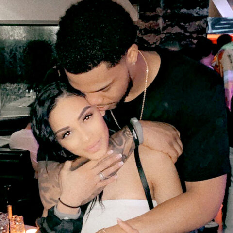 Couple embraces in a dim club; man kisses the woman's head as she looks at the camera, tattoos visible on her shoulder area.