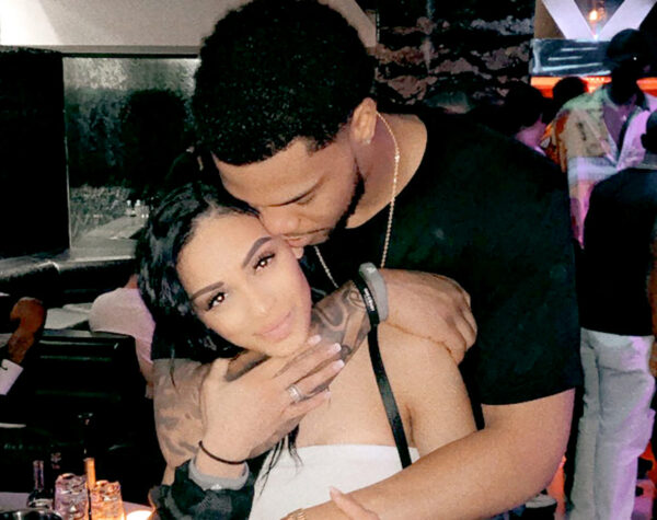 Couple embraces in a dim club; man kisses the woman's head as she looks at the camera, tattoos visible on her shoulder area.