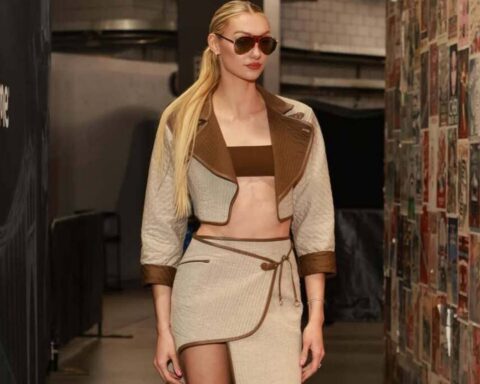 Model walking backstage in a beige and brown two-piece outfit with a cropped jacket and wrap skirt, wearing sunglasses.